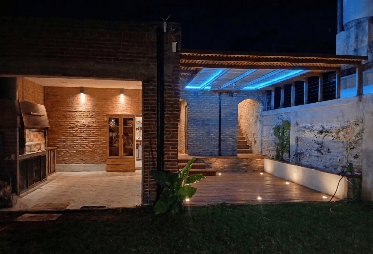 Quincho al fondo con deck y pergola de madera, sector de parrilla cubierta a la izquierda.
